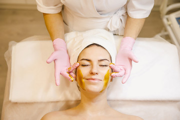 Hands of cosmetology specialist applying gold facial mask using brush, making skin hydrated and face glowing and skin. Attractive brunette relaxing with closed eyes and enjoying spa procedures