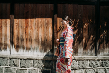side view of young local girl in pink floral kimono dress walking along the wooden wall under tree shadow. beautiful japanese woman in traditional cloth cardigan relaxing enjoy peaceful environment