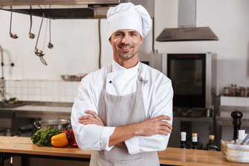 Image of caucasian male chief in white uniform smiling, while standing at kitchen in restaurant