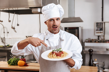 Photo of professional male chief in white uniform holding plate with pasta, while cooking at kitchen in restaurant