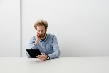 Thirty something bearded man looking thoughtfully on his tablet sitting at an empty light colored desk.