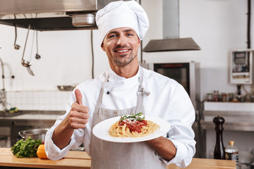 Photo of smiling male chief in white uniform holding plate with meal, while cooking at kitchen in restaurant