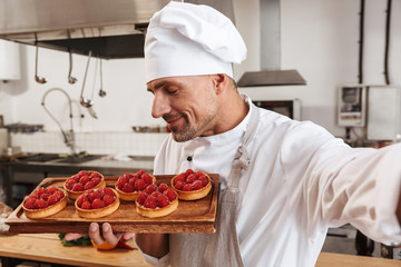 Photo of professional male chief in white uniform taking selfie and holding plate with cakes, while cooking at kitchen in restaurant