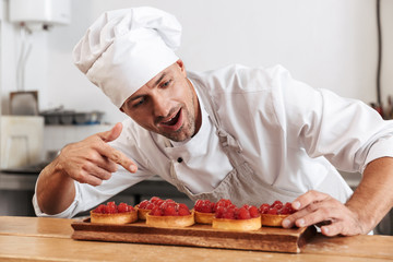 Photo of pleased male chief in white uniform holding plate with cakes, while cooking at kitchen in...