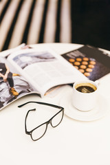 Cropped vertical photo of white table with some magazines, cup of coffee, selective focus on glasses/ energy charge in the morning, delicious hot black coffee at table, workplace, coffee mug.