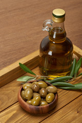 wooden tray with oil bottle, bowl of olives and olive tree leaves on brown surface