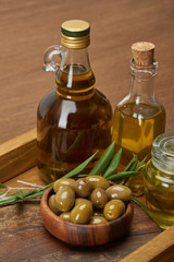 wooden tray with different oil bottles, bowl of olives and olive tree leaves on brown surface