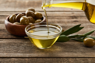pouring oil from bottle into glass bowl, bowl of olives, olive tree branch and olive on wooden...