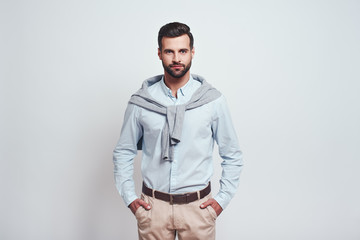 Fototapeta premium Irresistible man. Bearded young man is looking at camera, keeping hands in his pockets while standing against grey background