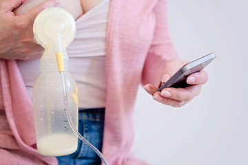 Mother is using single electric breast milk pump in one hand and phone in another one. - image