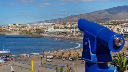 Blue coin operated binoculars pointing towards Playa Fanabe, in Las Americas resort, with tourists enjoying vacation and leisure, Costa Adeje, Tenerife, Spain © Ana