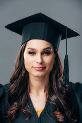 smiling indian student in academic gown and graduation cap, isolated on grey
