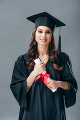 cheerful indian woman in academic gown and graduation hat holding diploma, isolated on grey