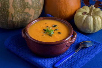 Pumpkin vegan soup in a clay bowl served with parsley, olive oil and pumpkin seeds on the blue background