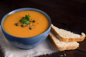 Pumpkin vegetarian soup in a blue bowl served with parsley, olive oil and pumpkin seeds on a dark wooden background.