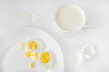Very light photo with a predominance of white. On a white background there is a plate with sliced boiled eggs and a cup with milk.