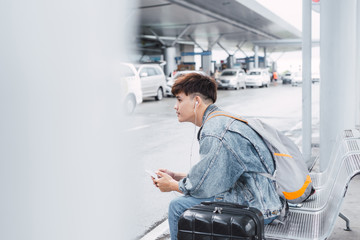 young man is sitting and waiting for bus. He is holding backpack and listening to music from earphones. Guy is looking forward with aspiration