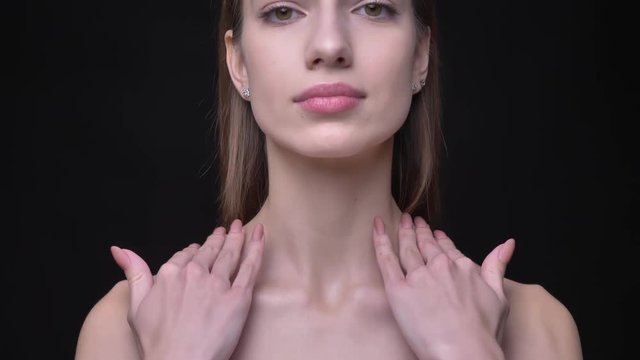 Close-up portrait of young caucasian girl with nude make-up touching softly her collarbone on black background.