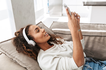 Naklejka premium Photo of modern african american woman wearing headphones holding cell phone, while lying on sofa in bright apartment