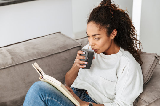Image of young african american girl reading book and drinking tea, while sitting on sofa in bright flat