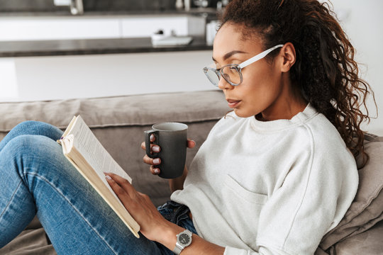 Image of smart african american girl reading book and drinking tea, while sitting on sofa in bright flat