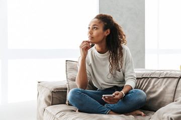 Photo of pretty african american woman using cell phone, while sitting on couch in bright apartment