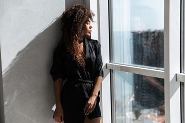Portrait of brunette african american woman wearing housecoat, standing near window indoor