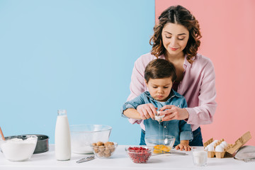 smiling mother with concentrated little son cooking together at white kitchen table on bicolor background