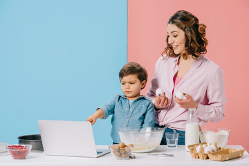 little boy pointing at laptop display while cooking with mother on bicolor background