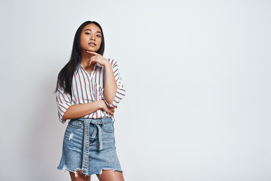 Elegance and beauty. Young asian woman in striped shirt with hand on chin looking at camera while standing against grey background