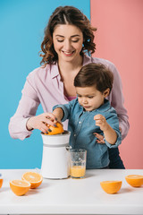 happy mother with adorable son squeezing fresh orange juice together on bicolor background