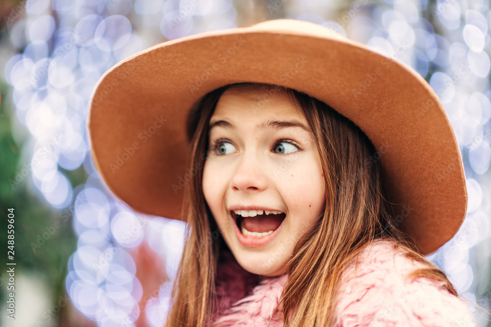 Wall mural outdoor portrait of pretty young kid girl wearing brown hat and pink coat, street fashion for childr