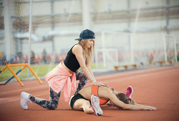 Young beautiful woman with blonde hair helps another woman to stretch her legs in the sports hall...