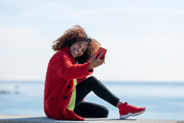 Side view of young beautiful curly african american woman sitting on a bench at beach while using a mobile phone outdoors
