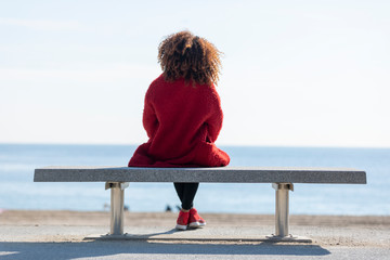 Rear view of a young curly woman wearing red denim jacket sitting on a bench while looking away to horizon over sea - Powered by Adobe