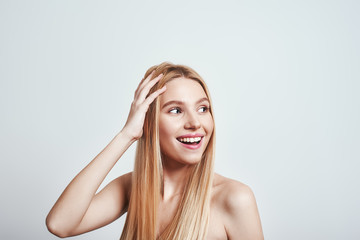 Naklejka premium Feeling free and happy. Attractive young woman with naked shoulders touching her hair, looking away and smiling in studio on a grey background