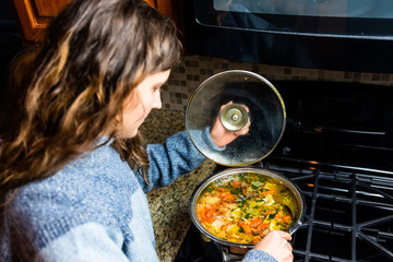 Large big pot of homemade vegetable soup and woman stirring on granite counter top in kitchen in...