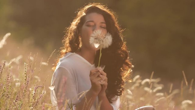 Beautiful young woman blows dandelion in a wheat field in the summer sunset. Beauty and summer concept