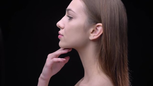 Portrait in profile of young and slim caucasian girl with nude make-up tenderly touching her neck on black background.