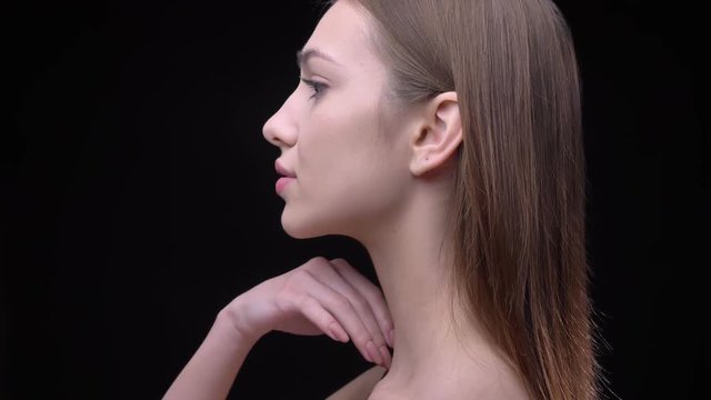 Portrait in profile of young and slim caucasian girl with nude make-up softly touching her neck on black background.