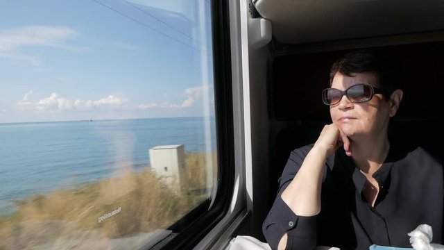 Senior woman in black shirt and sunglasses pensively sitting near window in train cabin, admiring bright sunny sea view. Slow motion