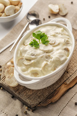Thick creamy sauce with mushrooms in a deep white sauce-pot stands on a wooden board. High key.