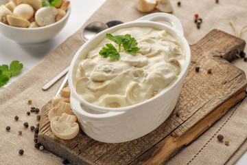 Thick creamy sauce with mushrooms in a deep white sauce-pot stands on a wooden board. High key.