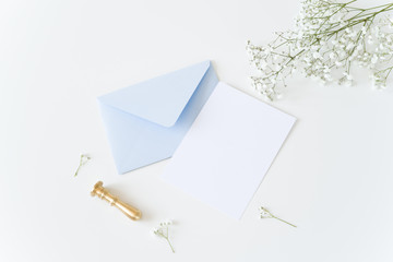 White and blue wedding stationery. Blank greeting card, craft envelope, washi tape and golden stamp, binder clips with olive branch.White table background. Flat lay, top view.