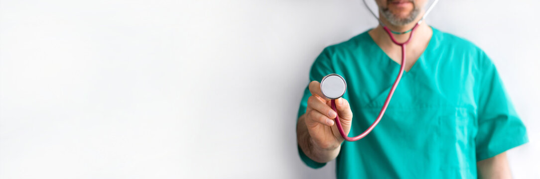 defocused doctor with beard in green uniform with stetoscope in hand in focus ready to examin on white background with copy space. Horizontal long banner, medicine, cardiology, heart checkup