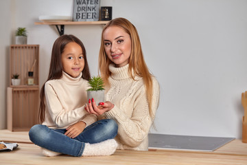 Beautiful mom and her small little daughter on weekend, sitting on countertop, consider a flower in...