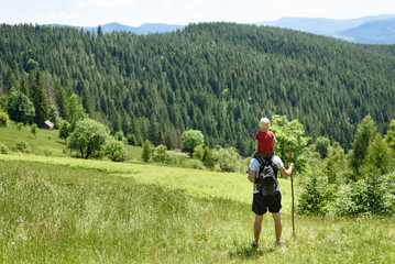 Father with son on his shoulders standing with staff in the background of green forest, mountains and sky with clouds. Back view