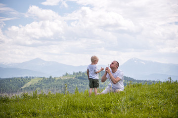 Fototapeta na wymiar Father and young son are blowing dandelions sitting in the grass on a background of green forest, mountains and sky with clouds. Friendship concept