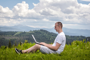 Naklejka na ściany i meble Man with laptop sitting on green grass on a background of mountains. Side view