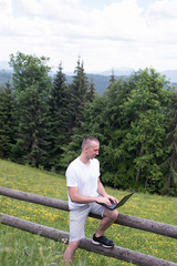 man sits on a wooden fence and works with a laptop near the field and coniferous forest. Vertical frame